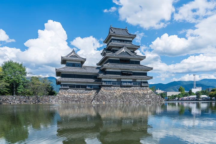 A serene scene features a large, multi-level castle-like structure on a stone pedestal, surrounded by a stone wall and a serene body of water. The sky is a clear blue with white clouds, and mountains are visible in the distance. The castle's reflection is visible on the water, creating a mirror-like effect.