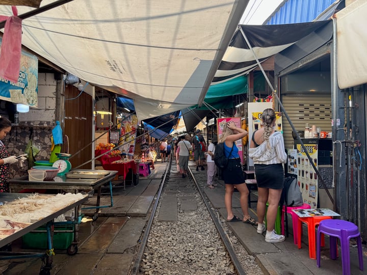 A bustling market in a developing country features colorful stalls, people shopping, and a train track in the background.