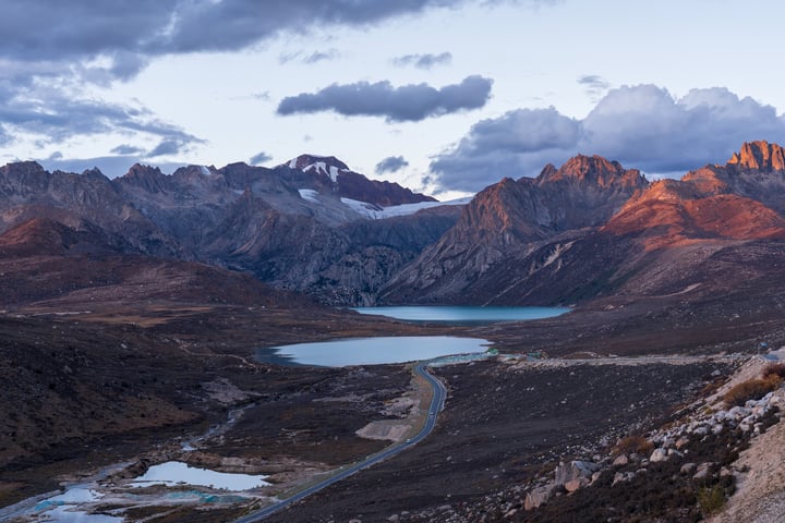 A breathtaking mountain landscape at sunrise or sunset, with a serene blue lake reflecting the sky, surrounded by rugged mountains and a winding road.