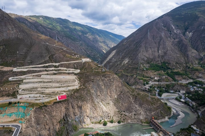 An aerial view of a mountainous valley with steep cliffs, a winding river, and a bridge, surrounded by mountains and greenery.