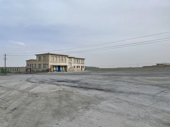 An old, dilapidated building sits in a barren, empty lot, surrounded by a fence and a solitary car.