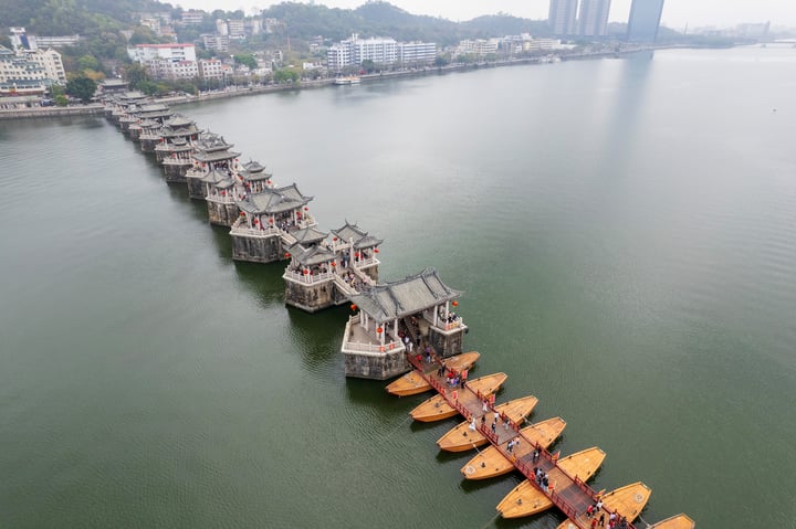 这是一座横跨河流的古桥，桥墩上建有多座传统风格的亭阁，河面上有数艘黄色小船，背景是城市建筑和远处的山脉。