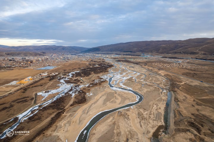 An aerial view of a river winding through a valley, surrounded by mountains and a city, with a cloudy sky above.