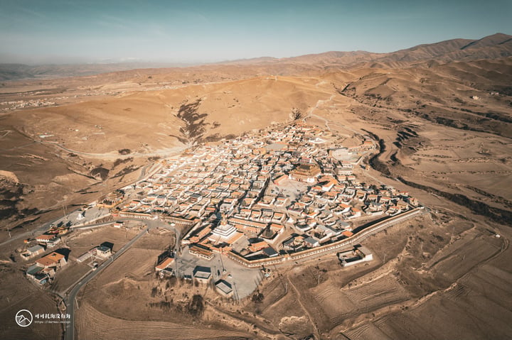 An aerial view of a town in a desert-like area, surrounded by mountains and fields, with a clear blue sky above.