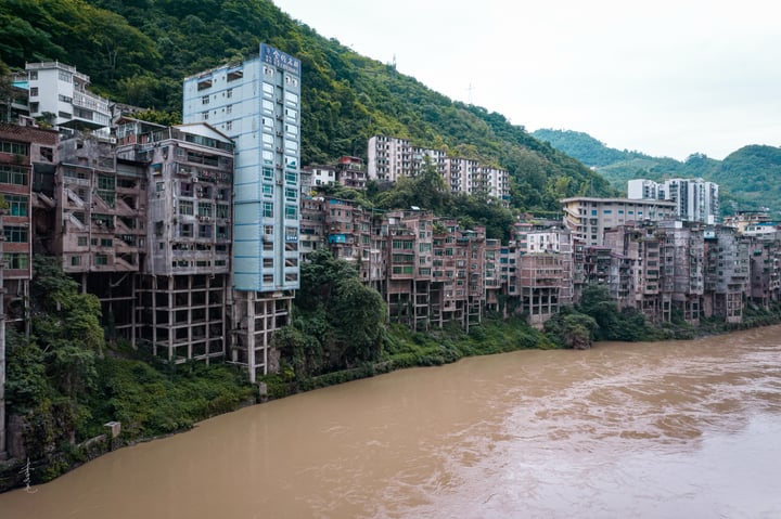 A cityscape on a hillside, with tall buildings of varying heights and colors, surrounded by lush greenery. The river, a brown, choppy body, flows through the city, reflecting the overcast sky.