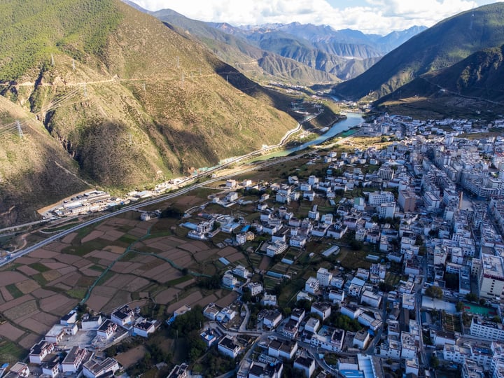 An aerial view of a mountainous valley with a river, surrounded by green fields and houses, with a clear blue sky above.