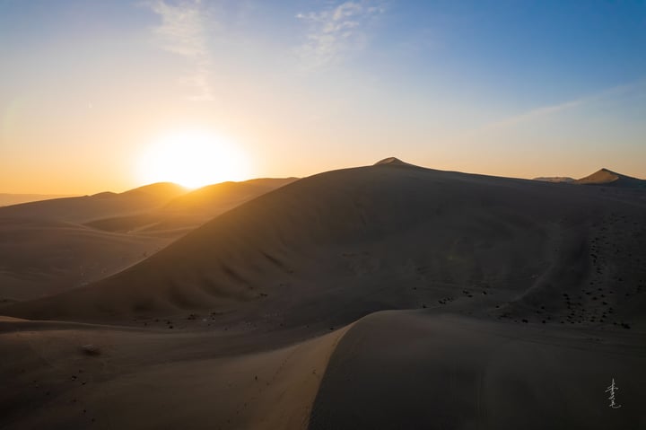 A serene desert scene at sunset, with a large sand dune in the foreground, a smaller one in the background, and a sun casting a warm glow on the landscape.