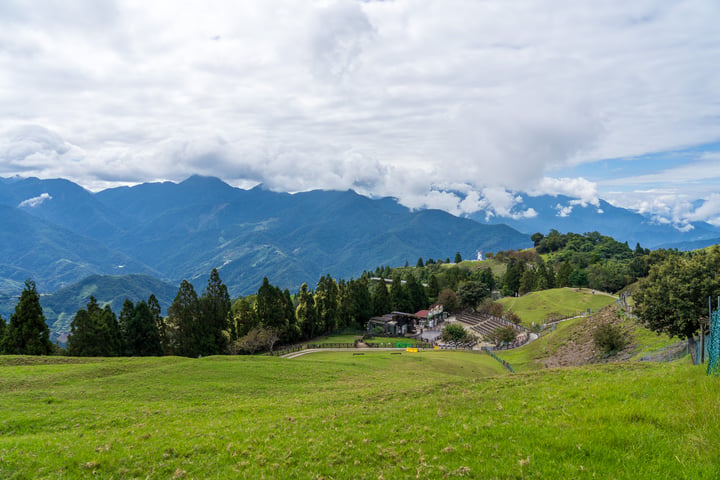 A serene mountain landscape with a lush green hillside, a small building, and a clear blue sky with white clouds.