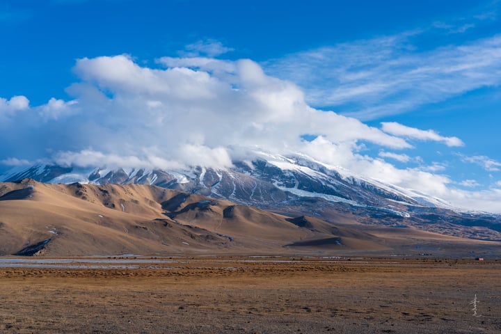 A vast, barren desert landscape with a snow-capped mountain in the background, a clear blue sky with scattered clouds, and a few distant figures on the left side.