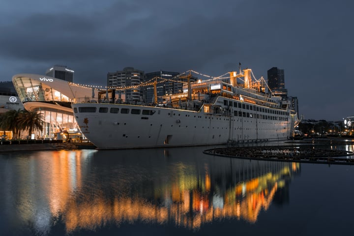 A large white cruise ship is docked in a calm body of water, with its reflection visible in the water. The ship is surrounded by modern buildings, including a large glass building and a tall structure with a curved roof. The sky is dark and cloudy, creating a serene atmosphere.