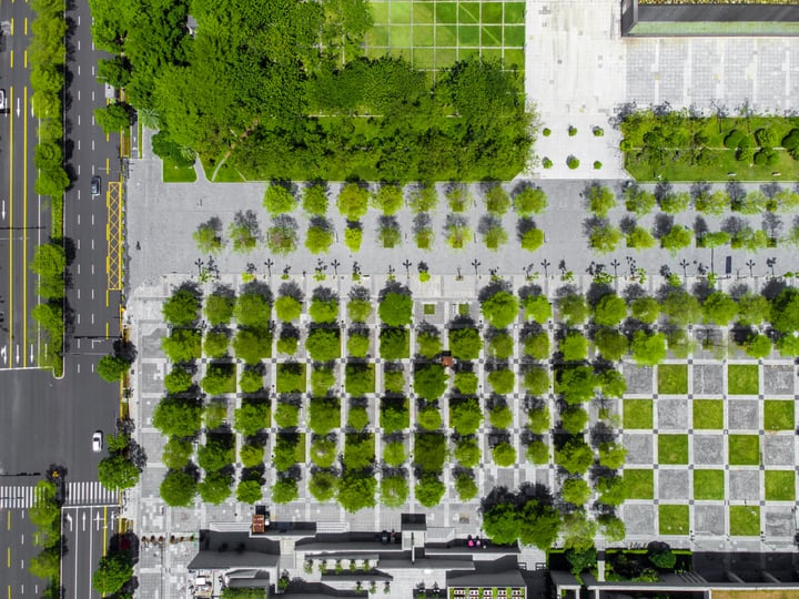 An aerial view of a city park with a grid of green spaces, surrounded by a road and buildings. The park is enclosed by a fence and has a sidewalk for pedestrians. The park is well-maintained and surrounded by trees and bushes. The road and buildings are visible in the background, and the park is the main focus of the image.