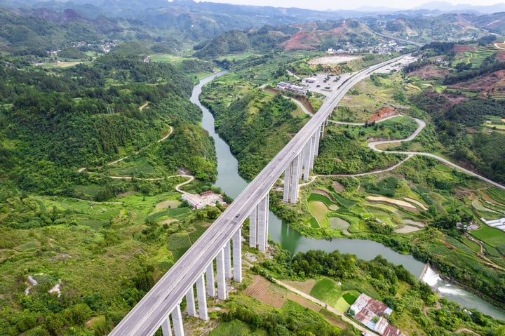 An aerial view of a long, white concrete bridge spanning a river in a valley, surrounded by lush green hills and valleys. The bridge is flanked by a winding road, and the river flows beneath it. The sky is clear blue, and the image is taken from above, showcasing the bridge's grandeur and the surrounding landscape.