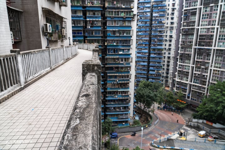 A high vantage point captures a bustling cityscape with tall apartment buildings, a busy street, and a park. The buildings are predominantly white and gray, with blue accents, and are adorned with air conditioning units. The street is filled with cars and a bus, and a park with trees and a fountain is visible in the background. The image is taken from a balcony overlooking the city, showcasing the urban environment.