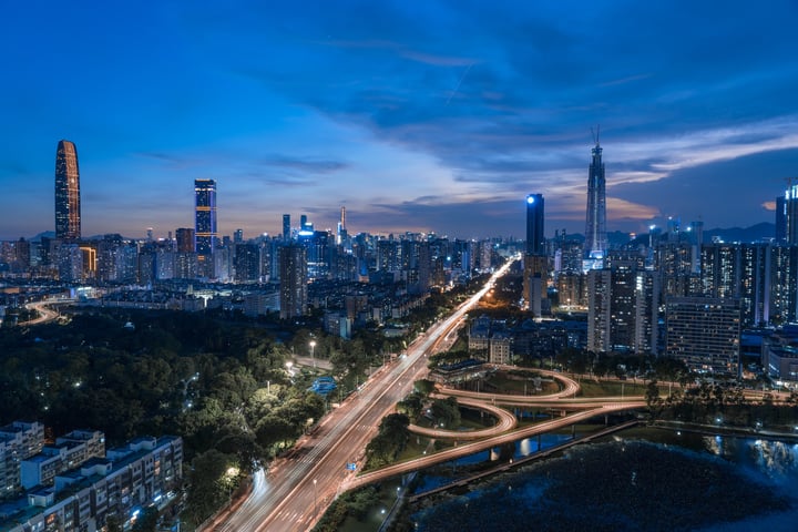 A nighttime cityscape features a tall, curved skyscraper with a curved top, a tall, narrow building with a flat top, and a tall, flat building with a curved top. The city is surrounded by a river and trees, with a dark blue sky above.
