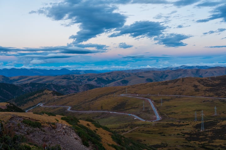 A winding mountain road winds through a valley, surrounded by lush green hills and mountains, with a cloudy sky overhead.