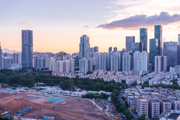 A cityscape under a sunset sky features tall buildings with varying heights and colors, including a prominent skyscraper. A construction site with a large red and white construction vehicle is in the foreground, surrounded by a mix of buildings and greenery. The scene is bathed in warm, orange and pink hues, creating a serene atmosphere.