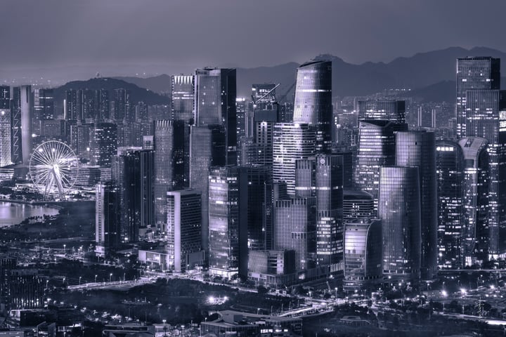 A black and white image captures a cityscape at night, with tall buildings illuminated against a dark sky. A Ferris wheel is visible in the distance, and mountains are visible in the background. The cityscape is densely populated, with lights illuminating the buildings.
