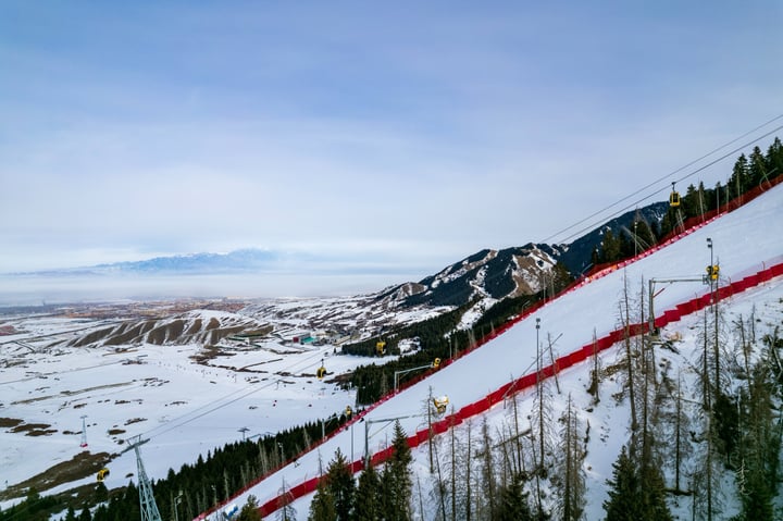 A snowy mountain slope with a red fence, a ski lift, and a vast landscape with snow-covered mountains and trees.