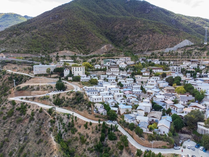 A picturesque mountain town with white buildings and blue roofs, surrounded by greenery and a winding road, is captured from an aerial perspective.