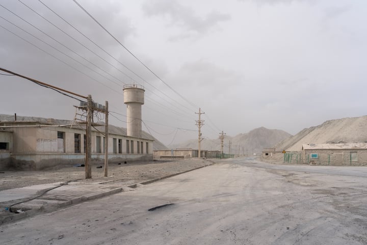 An empty, dusty road stretches into the distance, with a tall water tower and a small building on the left, and a large, empty building on the right. The sky is cloudy, and mountains are visible in the distance.