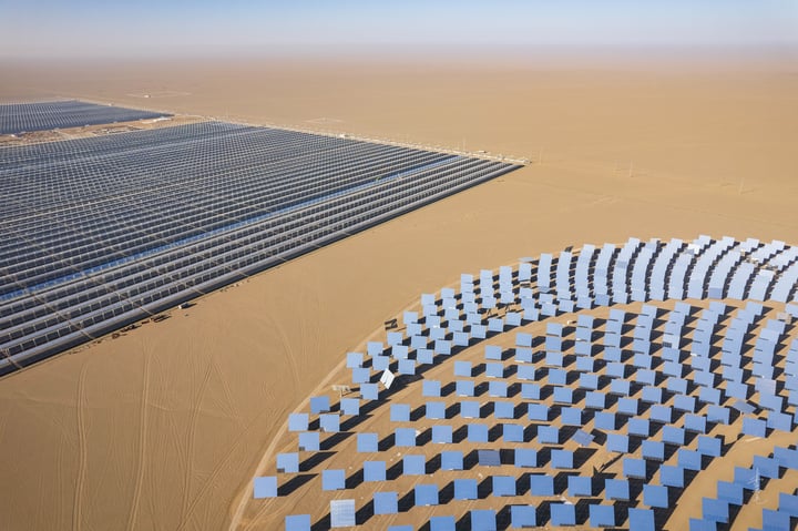 An aerial view of a large solar farm in a desert-like environment, with solar panels arranged in a circular pattern and a large white sun tower in the center. The farm is surrounded by a vast expanse of sandy terrain and a few wind turbines in the distance.