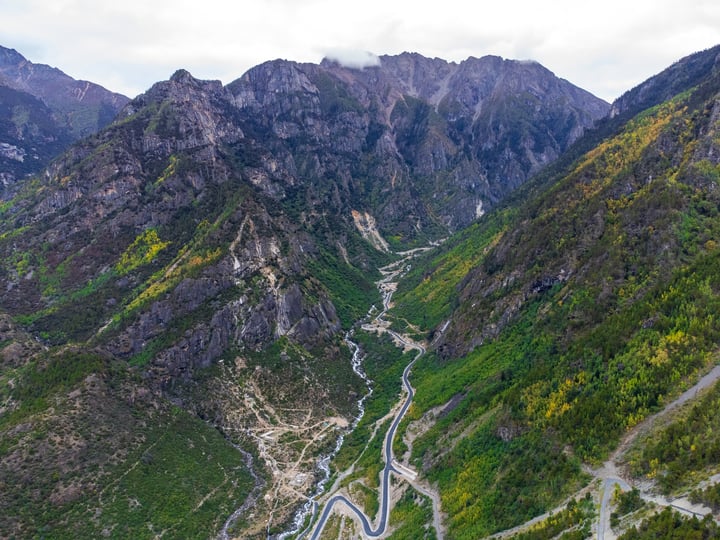 An aerial view of a winding mountain road through a valley, surrounded by lush green mountains and trees, with a cloudy sky above.