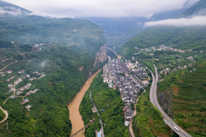 An aerial view of a valley with a winding river, surrounded by lush green mountains and a cloudy sky. The town is densely populated with white and gray buildings, and a highway runs through the valley.