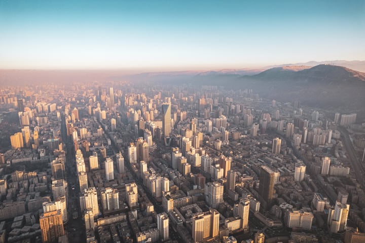 An aerial view of a bustling city with tall buildings, a mountain range in the background, and a warm, golden hue from the setting sun.