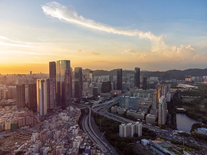 An aerial view of a bustling city at sunset, with tall buildings and a river in the foreground, surrounded by mountains.