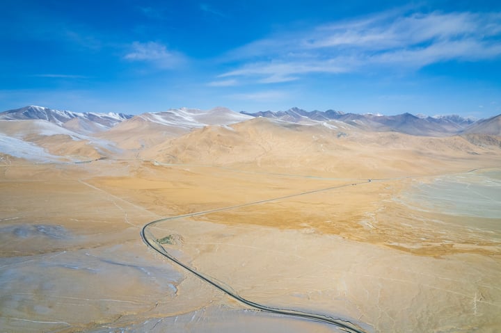 An aerial view of a vast, sandy landscape with a winding, curved road cutting through it. The mountains in the background are covered in snow, contrasting with the warm tones of the road. The sky is a clear blue with a few clouds, and the image is taken from a high vantage point, offering a panoramic view of the landscape.