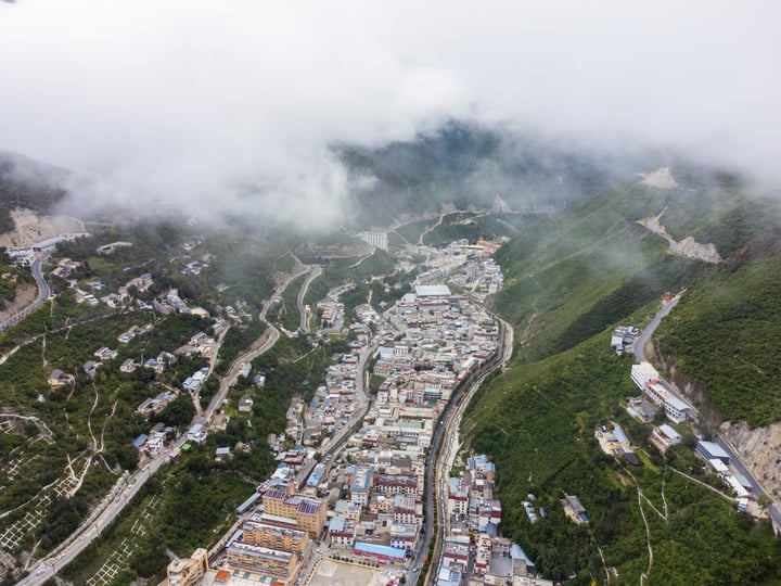 An aerial view of a Deqin County, surrounded by lush greenery and surrounded by mountains. The town is densely populated with houses and buildings, and the roads crisscross the valley. The town is surrounded by trees and hills, creating a serene and picturesque scene.