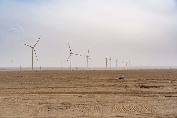 A desert landscape with a row of white wind turbines, a small vehicle, and tire tracks, under a cloudy sky.