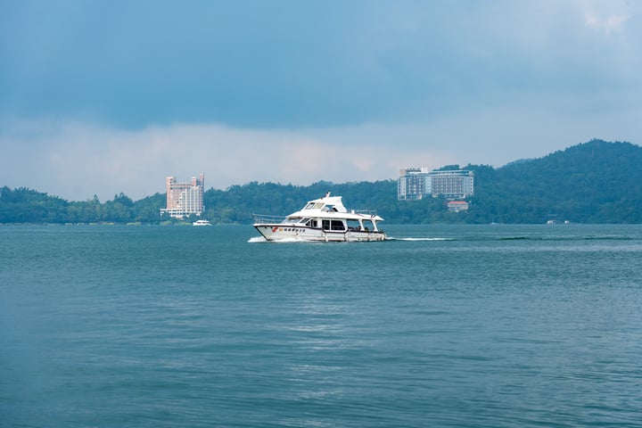 A white boat with a red stripe sails on a dark blue body of water, leaving a wake behind. In the distance, a city skyline with tall buildings and a mountain range is visible.