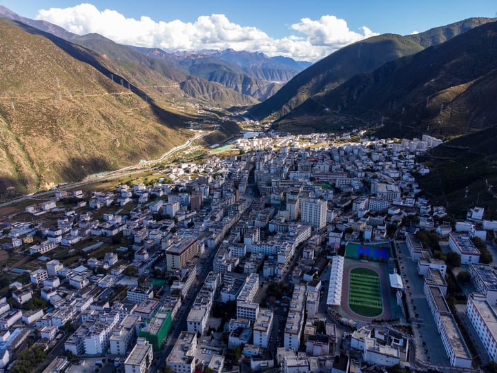 An aerial view of a mountainous city with white buildings, green fields, and a river, surrounded by mountains and a clear blue sky.