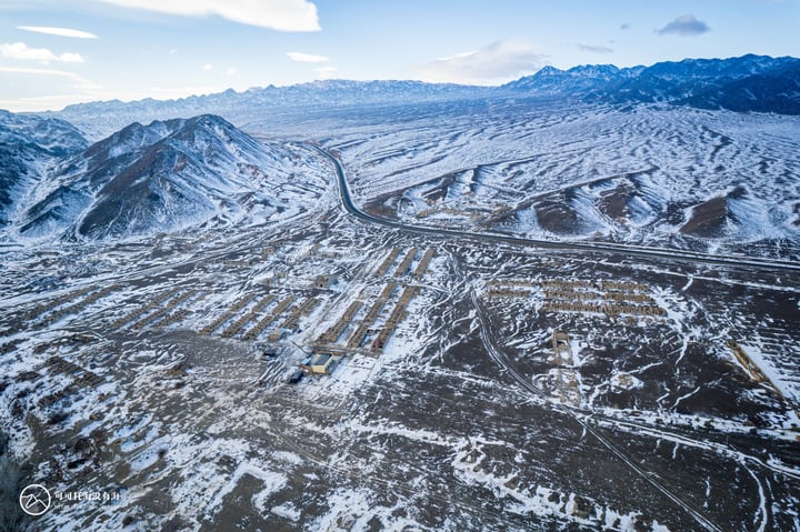 An aerial view of a snowy mountain landscape with a winding road, a small yellow building, and a few scattered trees.