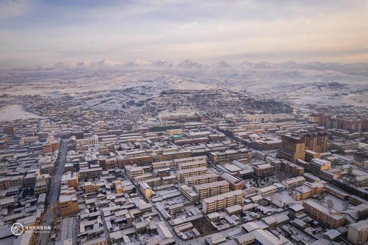 An aerial view of a snow-covered city with a mix of buildings and roads, surrounded by snow-covered mountains and a pale blue sky.