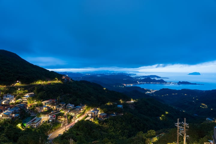 A serene night scene in a mountainous area, with a mountain range, a large body of water, and a hillside with houses and buildings illuminated by street lights.