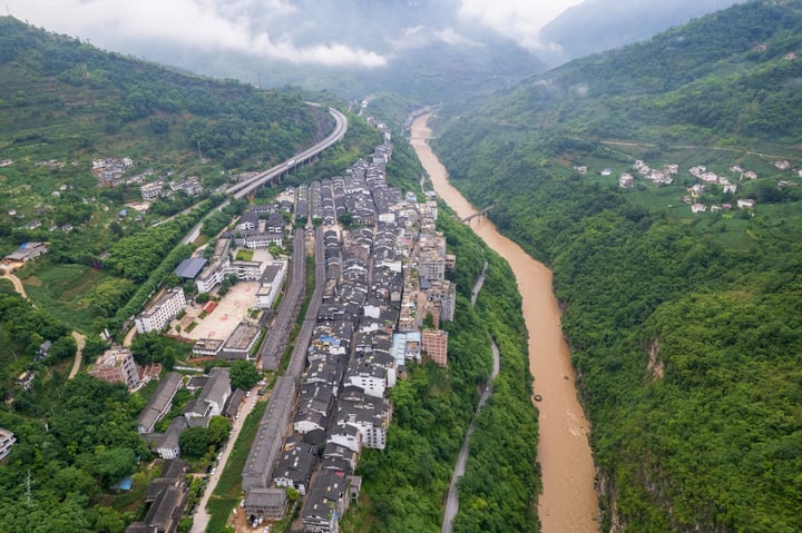 An aerial view of a city nestled between a river and mountains, with buildings of varying colors and shapes, surrounded by lush greenery. The river runs through the city, and roads crisscross the landscape. The sky is filled with clouds, casting a soft light over the scene.