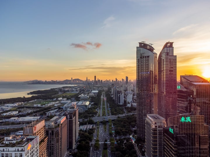A city skyline at sunset, with tall buildings and a road lined with trees, overlooking a body of water and a mountain range.