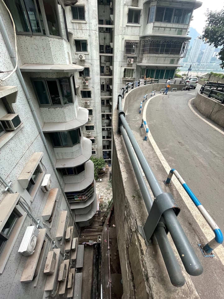 A city street with a curved concrete road, a tall white apartment building with air conditioning units, and a pedestrian walkway.