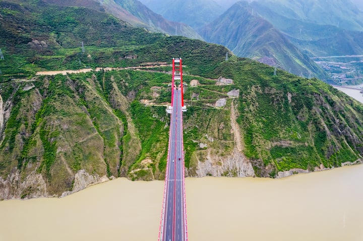 A red and white suspension bridge spans a river, with a white cable connecting the two sides. The bridge is surrounded by mountains and green hills, and the sky is cloudy.