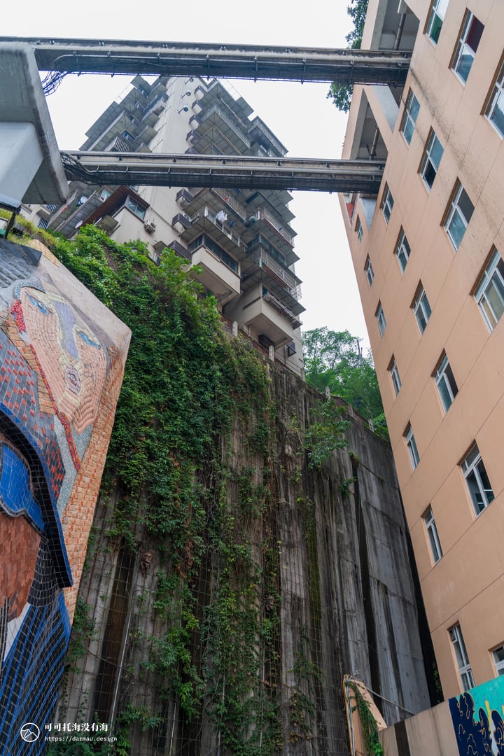 A cityscape features a tall, multi-level building with a unique, graffiti-covered wall, a beige building with a balcony, and a tall, white building with a balcony. The sky is gray, and the ground is covered in greenery.