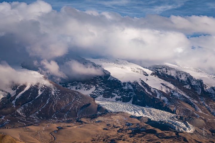 A breathtaking aerial view of a snow-covered mountain range with a large glacier, surrounded by a cloudy sky.