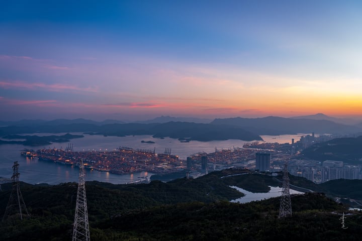 A cityscape with tall buildings, a harbor, and a bridge, surrounded by mountains and trees, bathed in warm hues of sunset.