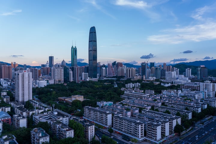 The image captures a cityscape at dusk, with a tall skyscraper dominating the skyline. The city is densely populated with buildings of varying heights, some with pointed tops. The sky is a deep blue with scattered clouds, and the sun is setting, casting a warm glow on the city. The city is surrounded by mountains, adding a natural element to the urban landscape.