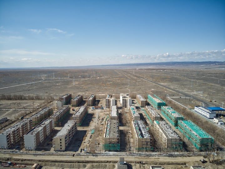 An aerial view of a large construction site with numerous buildings under construction, surrounded by a vast open field and a clear blue sky.