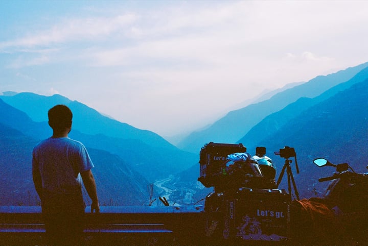A man stands on a ledge overlooking a valley, with a camera and tripod nearby, and a backpack on the ground.