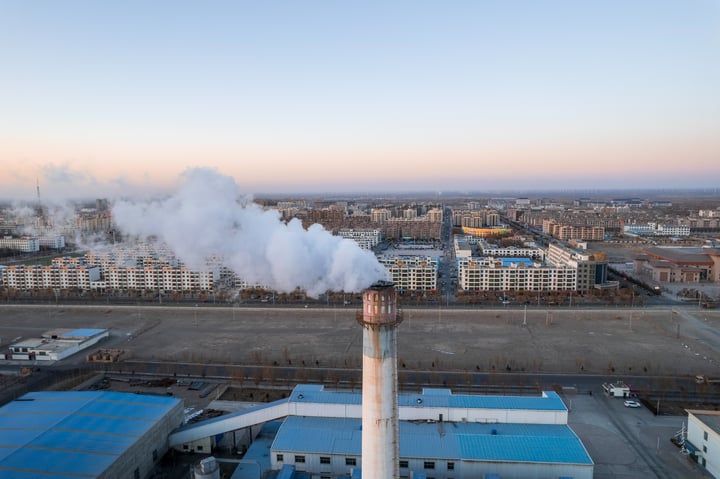 An aerial view of a large industrial complex with multiple buildings, including a large factory with smokestacks and a large factory with a large factory. The complex is surrounded by a large city with buildings of varying heights and colors. The sky is clear with a few clouds.