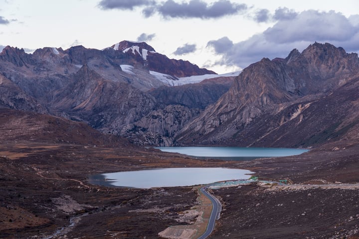 A serene mountain landscape with a blue lake, a winding road, and snow-capped mountains in the background.