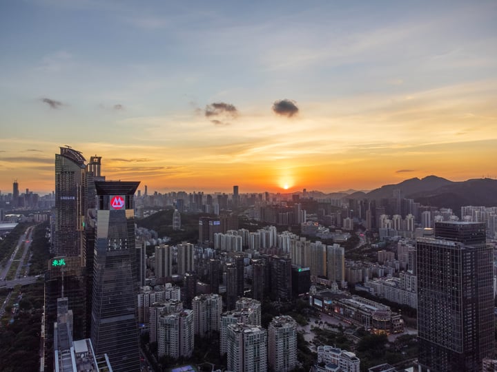 A city skyline at sunset, with tall buildings and a prominent skyscraper, reflecting the setting sun.